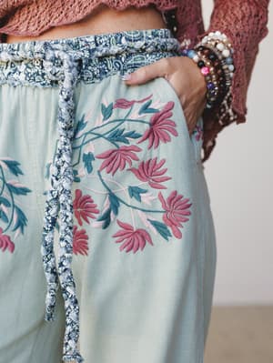 A close-up of sage wide-leg pants with lush floral embroidery in dusty rose and teal, paired with a mauve crochet top and stacked bead bracelets — the kind of outfit that looks like it has a good story behind it.