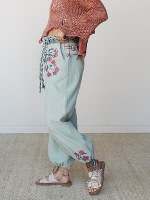 A side-profile shot of flowy sage green jogger-style pants with hand-painted pink floral embellishments and symbolic detailing at the waist, paired with a terracotta crochet top, floral tie anklets, and birkenstock-style sandals — the kind of outfit