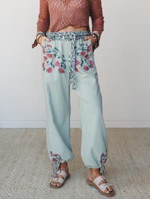 Soft sage harem-style pants with bold floral embroidery at the hip and ankles, styled with a rust crochet crop top, stacked bracelets, and leather sandals — the kind of outfit that looks like it was put together effortlessly but absolutely wasn't.