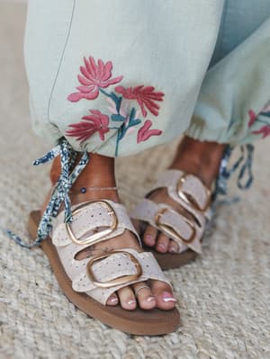 A close-up detail shot of embroidered sage linen pants with ribbon tie ankles, paired with blush double-buckle sandals on a jute rug — the kind of outfit that makes a farmers market feel like an event.