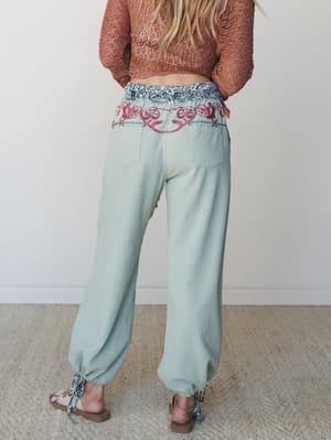 Shot from behind, a model wears relaxed sage green pants with bold floral embroidery along the waistband, paired with a terracotta crochet crop top and strappy sandals — earthy, expressive, and entirely her own.