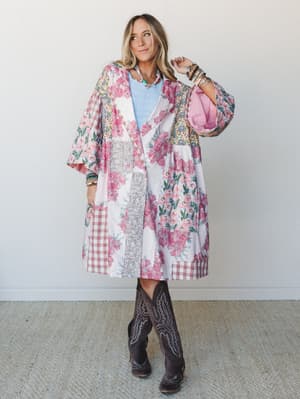 A long patchwork duster coat in soft pink florals, plaid, and ditsy prints is layered over a light blue knit top and worn with dark brown cowboy boots — the kind of outfit that looks like it was pulled from a very stylish grandmother's wardrobe and m