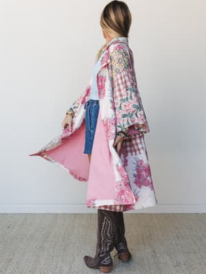 A flowy maxi-length patchwork jacket in pink florals and plaid opens wide to reveal a soft pink lining, styled over denim shorts and a ruched top with brown cowboy boots — effortlessly boho with a little Western heart.