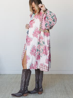 A long floral patchwork jacket in soft pink and cream layers over a denim short set, styled with tall dark brown cowboy boots and layered beaded jewelry — the kind of outfit that feels equally at home at a concert or a weekend farmer's market.