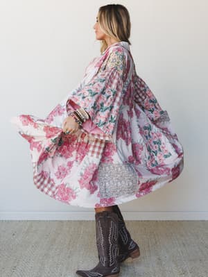 A flowy patchwork duster jacket swirls mid-twirl against a clean white wall, styled with dark cowboy boots and stacked bracelets — the kind of piece that makes you spin just to see what it does.