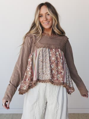 A mixed-fabric boho top with long knit sleeves and a flowy patchwork front — florals, stripes, and lace trim all living together in the best way — paired with white wide-leg lace-trim pants against a clean studio backdrop.