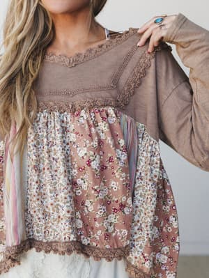 A close-up shot of a mixed-fabric babydoll top in warm tan, featuring patchwork floral panels and delicate crochet trim — the kind of top that looks like it was made from your grandmother's best vintage scarves, in the best way.