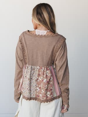 A back-view shot of a long-sleeve tan top with a mixed-media lower panel featuring patchwork floral and striped fabric, finished with crochet lace trim throughout — the kind of top that looks like it found you at a vintage market and never left.