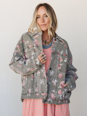 An oversized floral denim jacket in washed khaki layered over a pink striped dress, styled with silver stacked rings and a layered necklace — the kind of outfit that looks like it came together effortlessly but absolutely didn't.