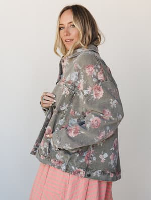 A model glances back over her shoulder in an oversized floral-print denim jacket in washed khaki, layered over a pink striped skirt — the kind of piece that makes a whole outfit feel intentional without trying too hard.