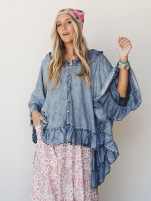 A flowy acid-washed denim poncho with dramatic ruffled sleeves and a button-front placket, styled over a pink floral maxi skirt with a embroidered head scarf and stacked bracelets — the kind of outfit that looks like it was assembled effortlessly but