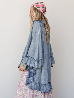 A smiling model shot from behind at a three-quarter angle, wearing a flowy acid-wash denim poncho with cascading ruffle tiers, layered over a pink floral midi skirt, topped with a pink crochet strawberry bandana — the whole look feels like a vintage