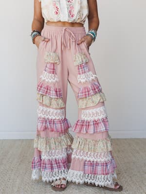 A dreamy, maximalist wide-leg pant in blush pink with tiered ruffles in mixed floral and plaid patches separated by lace trim, styled with an embroidered crop top and stacked beaded bracelets for a full boho moment.