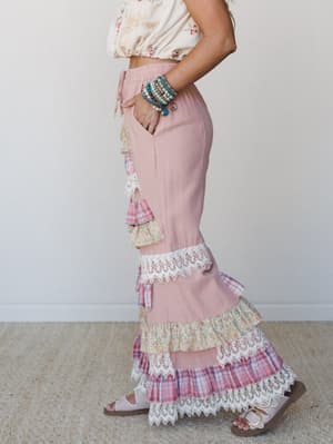A side-profile shot of tiered ruffle wide-leg pants in dusty pink, layered with mixed floral and plaid ruffles finished in lace trim — styled with a floral crop top, stacked boho bracelets, and buckle sandals for a free-spirited, feminine look.