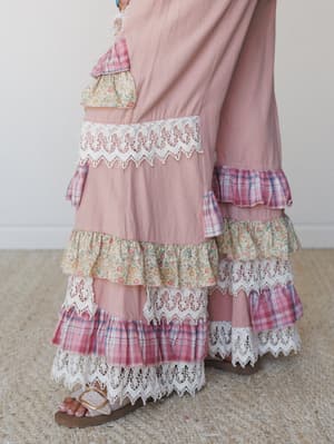 A close-up of wide-leg pants in dusty rose with alternating tiers of mixed-print ruffles — plaid, floral, and eyelet lace — layered all the way to the hem, styled with gold buckle sandals on a jute rug.