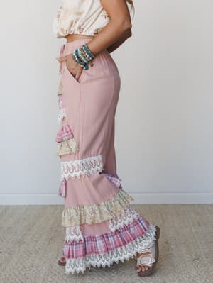 A side-profile shot of a tiered maxi pant in dusty rose, layered with mixed-print ruffles — plaid, floral, and lace trim — paired with a floral crop top and stacked turquoise bracelets for an effortlessly romantic boho look.