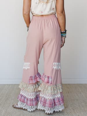 Shot from behind, these dusty pink wide-leg pants are the main event — tiered ruffle hems layered with plaid, floral, and lace trim give them a patchwork, vintage-fair energy that's equal parts whimsical and wearable.