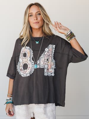 An oversized washed black graphic tee with lace-patched '84' appliqué is styled over a white eyelet skirt, layered with turquoise jewelry and stacked bracelets for an effortlessly boho, vintage-sporty feel.