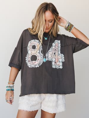 An oversized washed-black graphic tee with lace-trimmed '84' appliqué is styled with white eyelet shorts and layered turquoise jewelry — the kind of outfit that feels effortlessly thrown together but clearly wasn't.