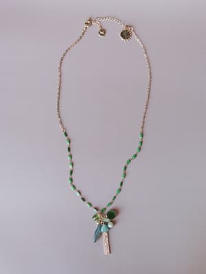 A delicate gold chain necklace with green enamel-detailed links leads down to a cluster pendant featuring mixed stones — deep green, pale mint, blue-grey, and white — alongside a textured gold bar charm, laid flat on a soft lavender-grey surface.