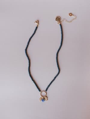A delicate dark navy crystal beaded necklace laid flat on a soft lavender-grey surface, with a gold ring pendant holding a cluster of small charms — a faceted blue stone, a hamsa hand, and a tiny gold charm — finished with a logo clasp and adjustable