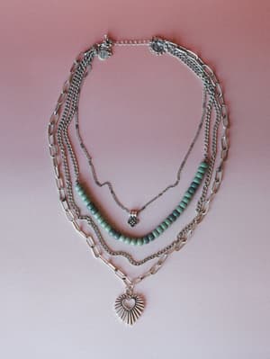 A layered silver necklace set arranged flat against a dusty pink background — mixing paper clip chains, a curb chain, turquoise stone beads, a floral charm, and a starburst heart pendant for that collected, worn-together look.
