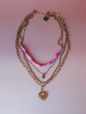 A layered gold necklace set laid flat against a soft pink background — mixing paperclip chain, curb chain, ombre pink beads, and a sunburst heart pendant for that perfectly collected, already-wearing-four-necklaces look.