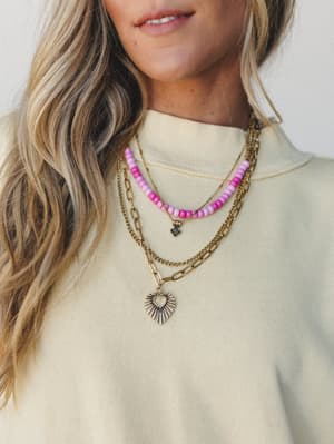 A close-up of layered gold necklaces styled together — a pink beaded strand with a small flower charm, a curb chain, and a paperclip chain with a sunburst heart pendant — worn over a cream mock-neck sweatshirt, feeling personal and collected.