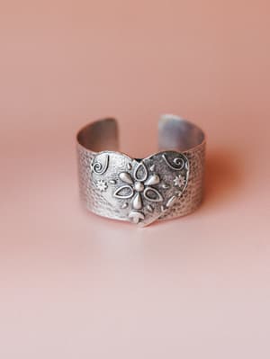 A wide silver cuff bracelet with an embossed heart centerpiece detailed with a raised flower motif, swirl accents, and hammered texture — shot against a soft blush background with that quiet, found-at-a-vintage-market kind of charm.