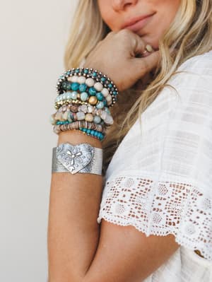 A close-up of a layered bracelet stack styled on a model in a white lace top — silver hammered heart cuff anchoring a mix of turquoise stone beads, cream and tan beads, gold accents, and a crystal-studded bangle, all with that collected-over-time fee