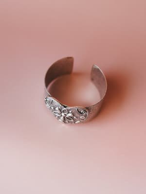 A wide silver cuff bracelet with an oxidized finish and embossed floral and scroll detailing, shot against a soft dusty rose background — the kind of piece that looks like it has a story.