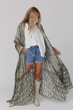 A woman wears the Paisley Tapestry Free Flow Duster Kimono in Olive over a white top and blue denim shorts, styled with bracelets, a necklace, and beige knee-high boots against a plain light background.