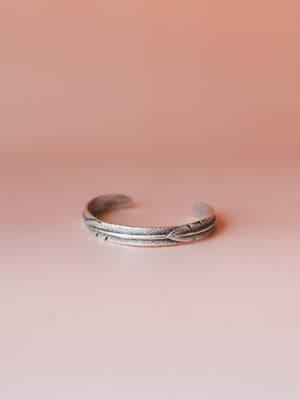 A silver cuff bracelet with an etched feather motif wraps open against a soft blush backdrop — the kind of piece that looks like it's been on your wrist forever.