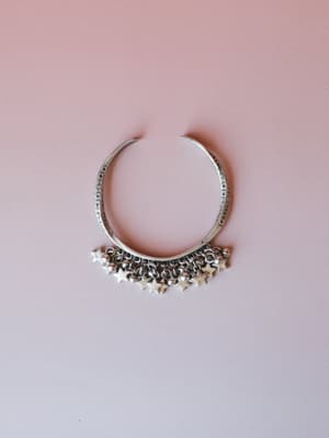 A silver-toned cuff bracelet with etched circle detailing along the band and a row of dangling star charms and small ball beads along the bottom edge, laid flat against a soft blush pink background.