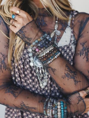 A close-up of layered boho jewelry styled on a model wearing a sheer navy lace overlay top — stacked bead bracelets, a chunky silver coin charm bracelet, and turquoise stone rings make this feel like a jewelry box came to life.