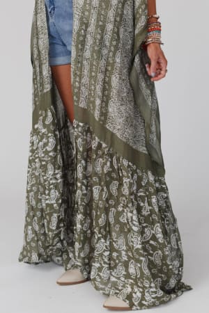 A person wearing blue denim shorts, tan pointed-toe shoes, multiple colorful bracelets on one wrist, and the Paisley Tapestry Free Flow Duster Kimono in Olive.