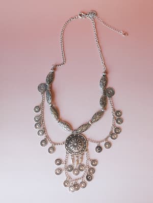 A layered silver statement necklace laid flat on a blush pink surface — engraved oval beads, a large floral medallion centerpiece, and rows of dangling coin charms give it that free-spirited, well-traveled energy.