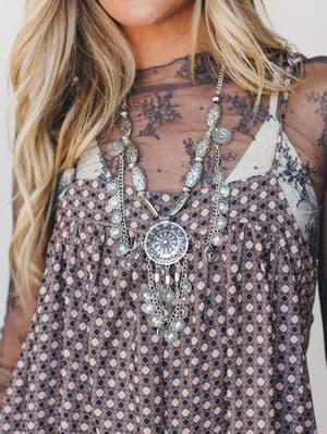 A close-up styled shot showcasing a statement silver medallion necklace with dangling coin charms, layered over a sheer floral mesh top and a diamond-print boho dress — the kind of outfit that makes someone stop and ask where you got everything.