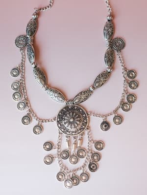 A statement silver bib necklace laid flat on a soft blush background — engraved oval links, a central mandala medallion, and cascading coin charms that feel like something you'd find at a market in Santorini and never take off.
