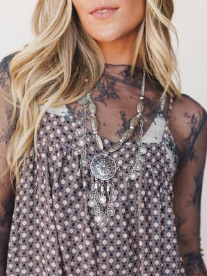 A close-up lifestyle shot of a layered boho look — a printed slip dress worn over a sheer lace top, styled with a long silver statement necklace featuring engraved coin pendants and chain fringe, giving off that free-spirited, effortlessly collected