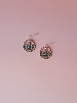 A pair of oxidized silver thunderbird drop earrings with turquoise stone centers, laid flat against a soft dusty rose background — earthy, storied, and the kind of thing you reach for every single day.