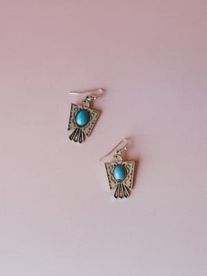 A pair of silver thunderbird drop earrings with turquoise stone centers and etched detailing, laid casually against a soft dusty pink background — the kind of earrings that make a simple outfit feel intentional.