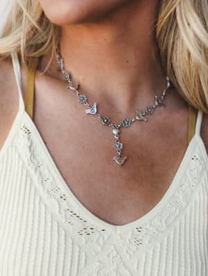 A silver charm necklace with bird and daisy motifs layered over a cream cable-knit tank — the kind of piece that feels like it belongs to someone with a good eye and a soft touch.