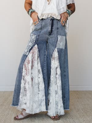 Wide-leg denim jeans with dramatic floral lace panel inserts sweep the floor while lace patchwork details at the hip add a vintage, handmade feel — styled with a white lace top and stacked boho bracelets for a full free-spirited moment.