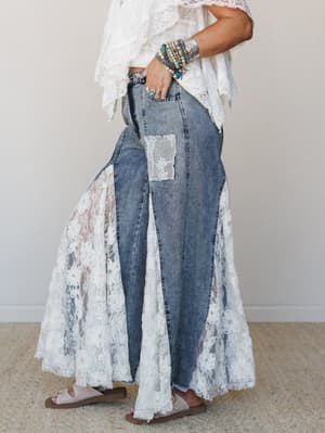 A side-profile shot of wide-leg denim jeans with dramatic floral lace panel inserts that flare out like a skirt, styled with a white lace-trim ruffle top, stacked turquoise bracelets, and flat sandals — the kind of outfit that makes people ask where