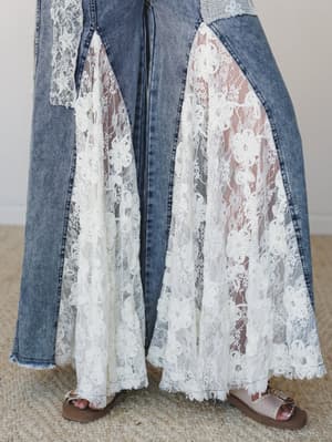 A close-up of wide-leg denim jeans with dramatic floral lace inset panels that flare out from the knee down, styled with strappy sandals on a jute rug — the kind of piece that makes you do a double-take in the best way.