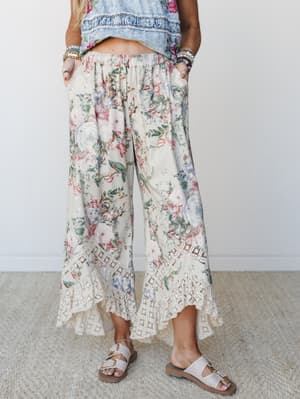 Wide-leg floral pants with a dreamy vintage garden print and lace hem trim, styled with layered bracelets and slide sandals against a clean white studio backdrop — the kind of pants that make you want to wander somewhere pretty.