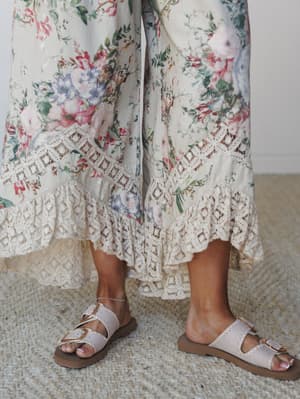 A close-up detail shot of wide-leg floral pants with a generous lace ruffle hem, paired with buckle slide sandals on a woven jute rug — soft, romantic, and effortlessly put-together.