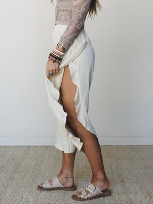 A side-profile shot of a cream linen wide-leg pant with a dramatic front slit, paired with a taupe crochet long-sleeve top, stacked bead bracelets, and buckle sandals — effortless and a little dreamy.