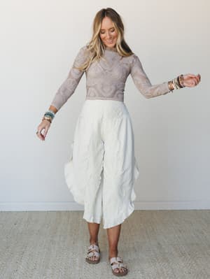 A smiling model twirls in a dreamy all-neutral pairing — a sheer lace long-sleeve crop top in taupe meets wide-leg textured white pants with a flowy, asymmetric drape, finished with beaded bracelets and birkenstock-style sandals.